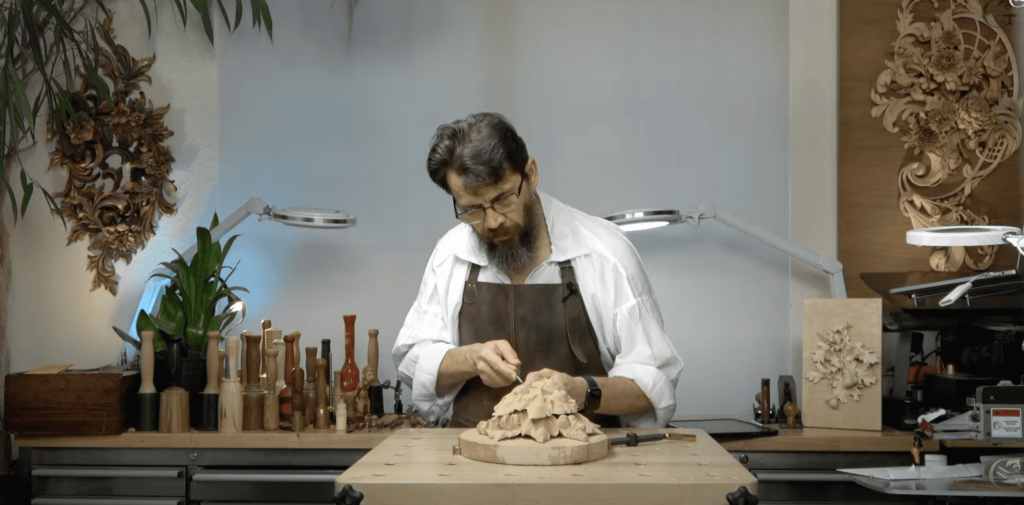 A woodworker is carving a detailed piece on a workbench filled with tools. The background shows a beautifully carved floral design, and there's a green plant to add a touch of life. You can see various carving tools neatly arranged, all set for some serious wood carving lessons. This scene is a perfect representation of what wood carving classes can look like—creative, focused, and full of inspiration!